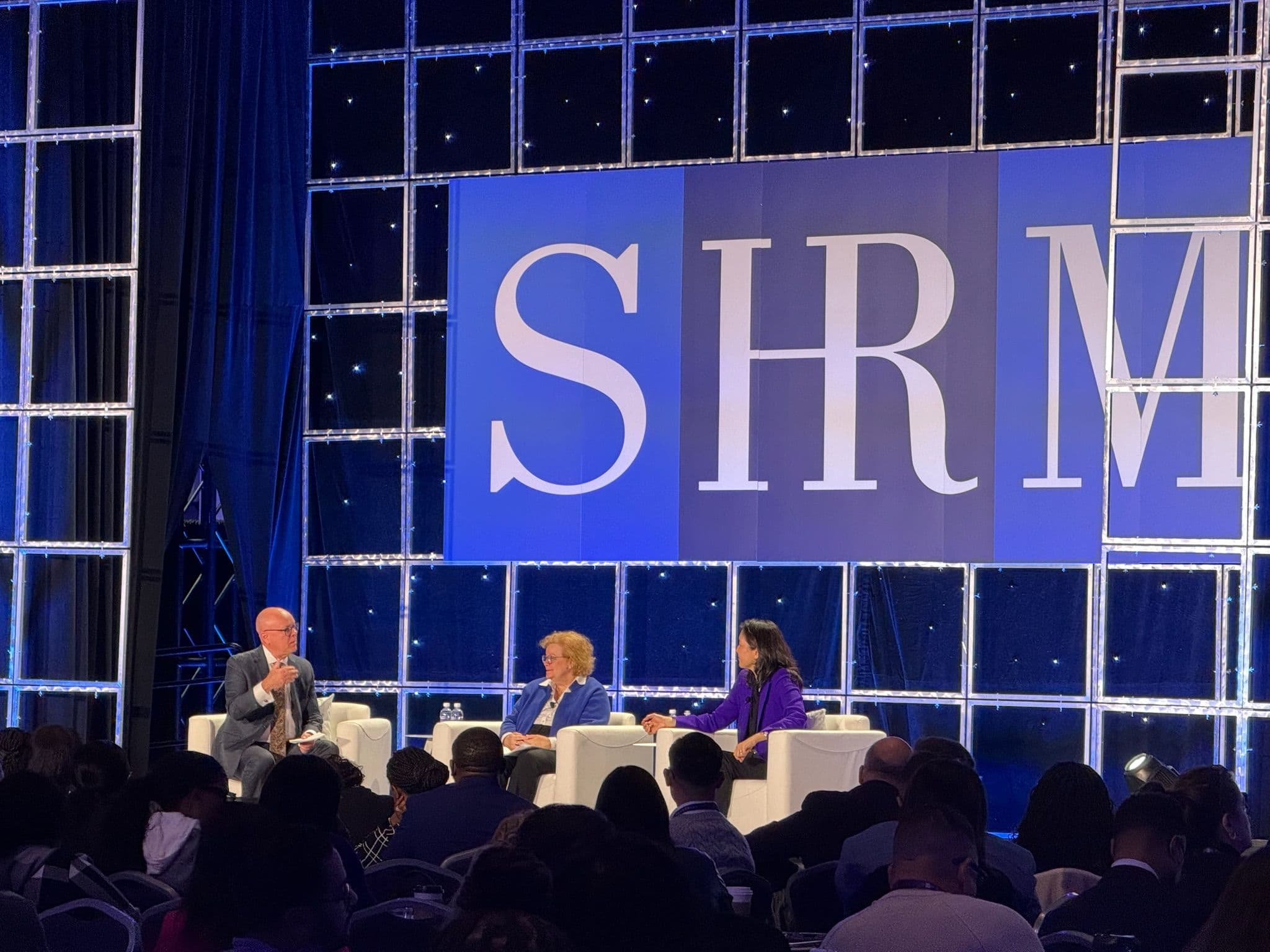 Panelists in white chairs on the SHRM main stage in front of a large SHRM logo, with a seated audience.